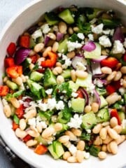 A large bowl of Mediterranean Bean Salad with serving utensils, lemon, and a jar of leftover vinaigrette off to the sides.