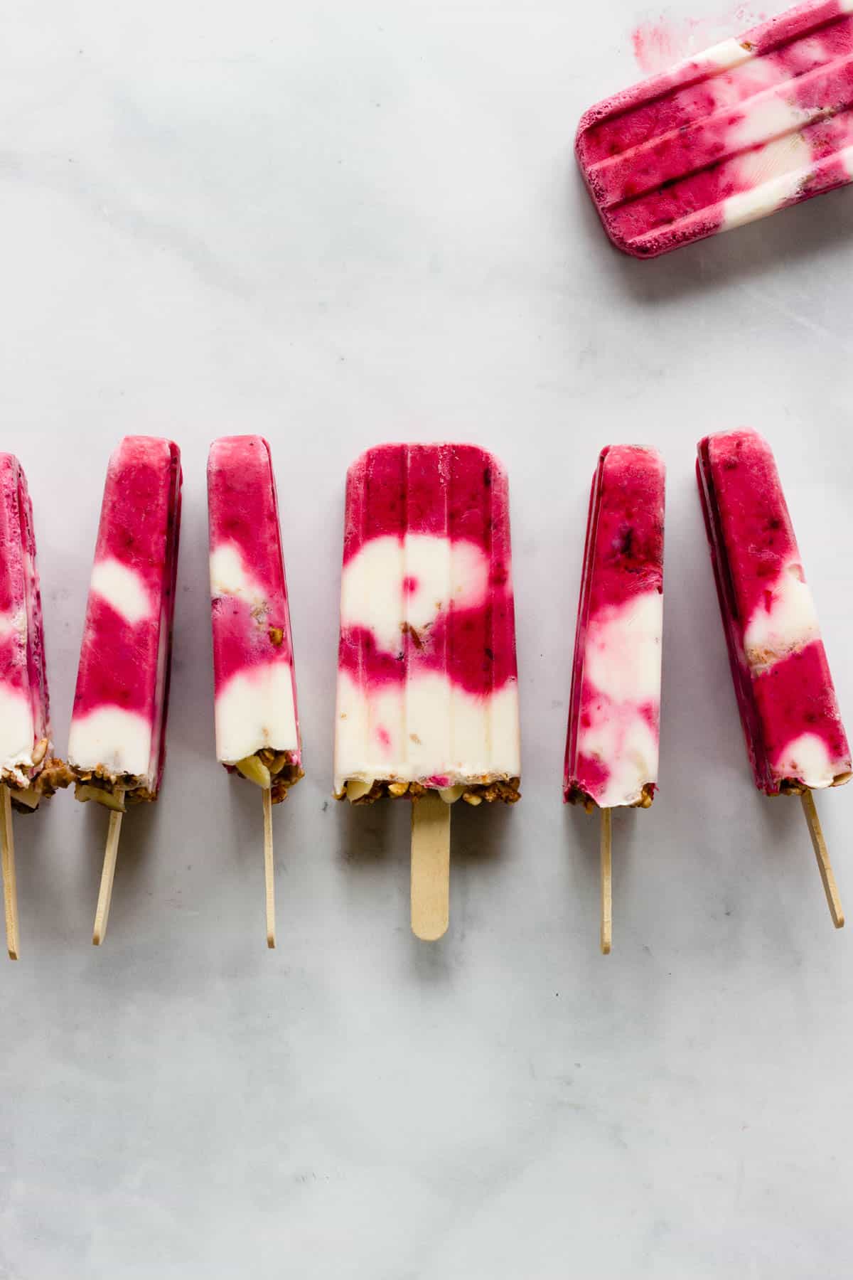 Berry yogurt popsicles are lined up horizontally on a marble countertop.