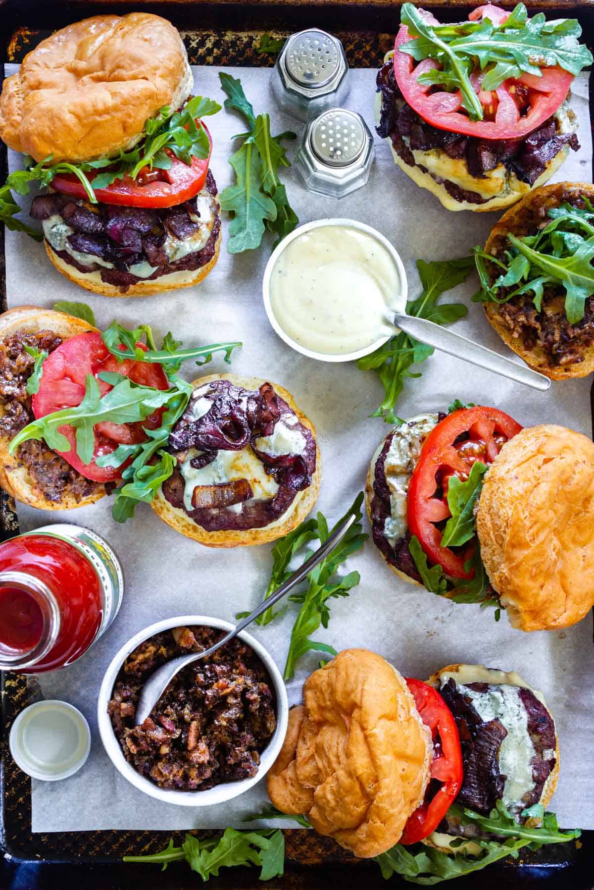 A zoomed in view of a tray loaded with bacon blue cheese burgers and small side dishes with roasted garlic aioli and bacon jam. A salt and pepper shaker and a ketchup bottle also accompany the burgers.