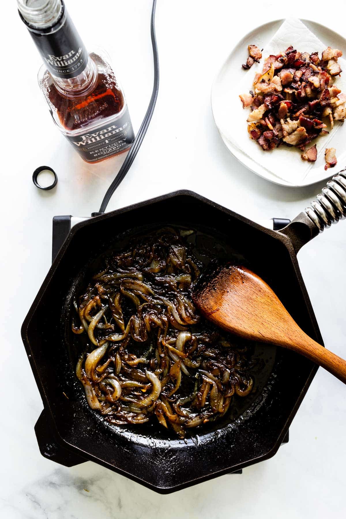 Sweet onions slowly caramelize in a cast iron skillet. A plate of cooked, coarsely chopped bacon and a bottle of bourbon sit nearby.