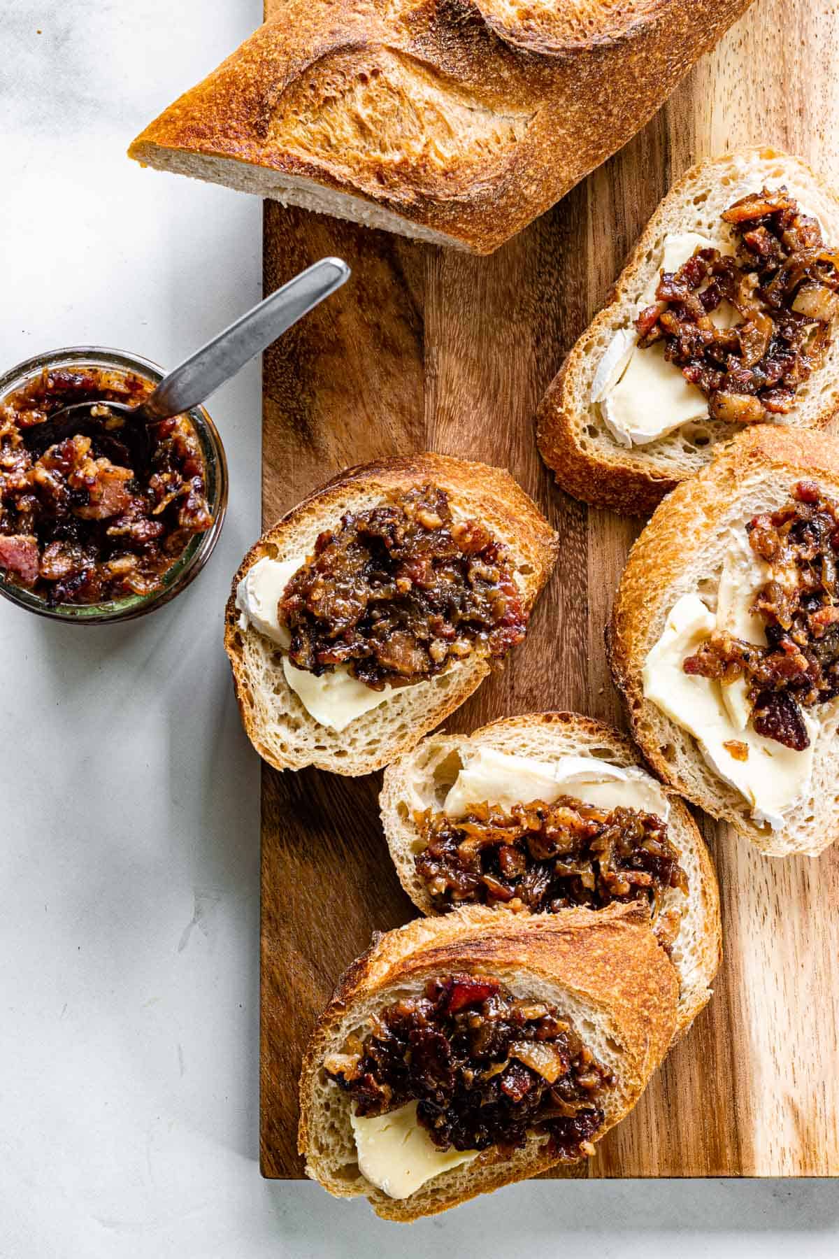 Crostini topped with gorgonzola and bacon jam are arranged on a wooden board. A jar of bacon jam with a spoon sits to the side.