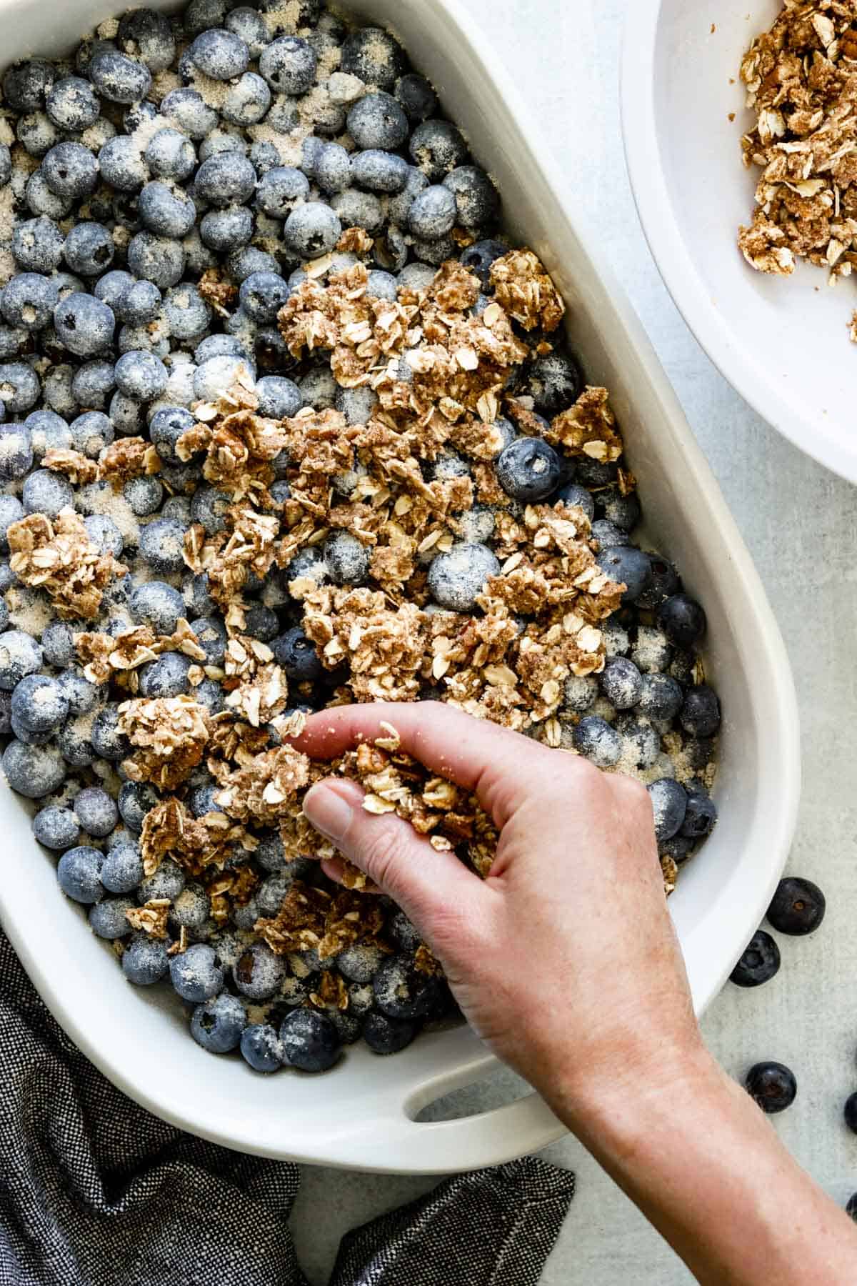 A hand is sprinkling crisp topping over a baking dish of blueberries.