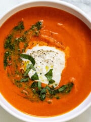 A white bowl of roasted red pepper tomato soup topped with burrata, basil, and a swirl of pesto.