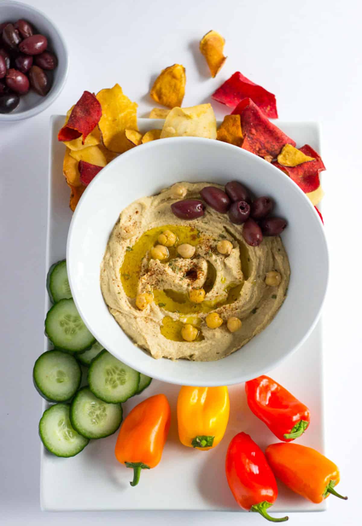 A bowl of smooth and creamy hummus surrounded by vegetable chips, mini bell peppers, cucumbers, and kalamata olives.