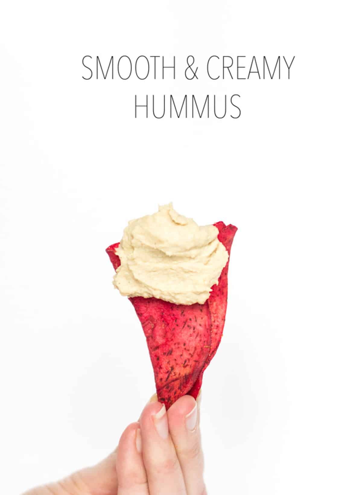 A hand holding a red vegetable chip with a swoop of hummus on top.