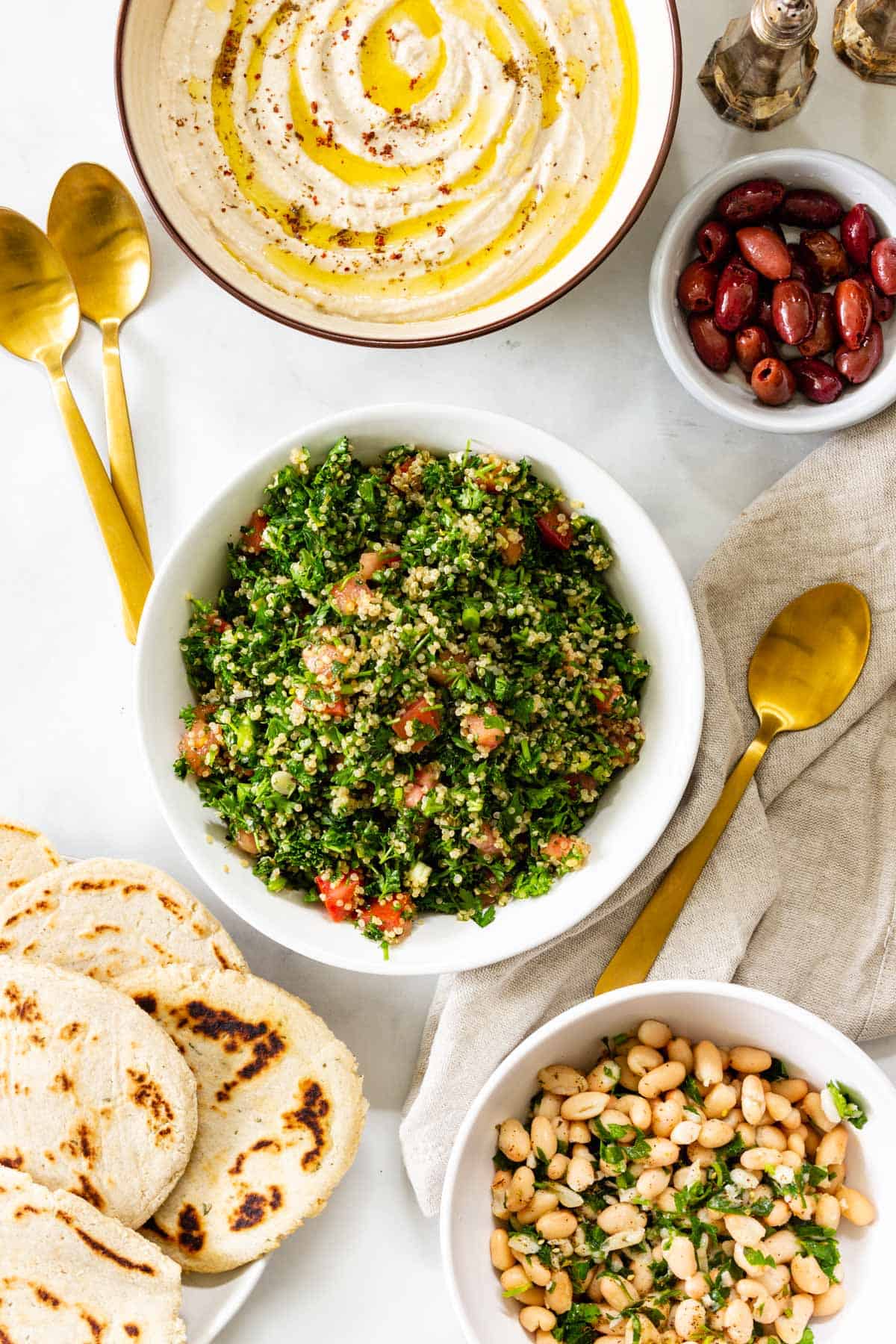 Plates of hummus, tabbouleh, olives, flatbread, and marinated beans are arranged on a table with gold silverware.
