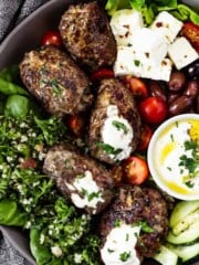 A large shallow bowl loaded with beef koftas, cucumber slices, tabbouleh, chunks of feta, olives, cheery tomatoes, and a dish of Greek yogurt sauce.