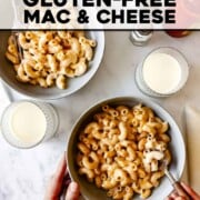 A table is set with two bowls of mac and cheese and cups of milk. Hands are holding a bowl and a fork.