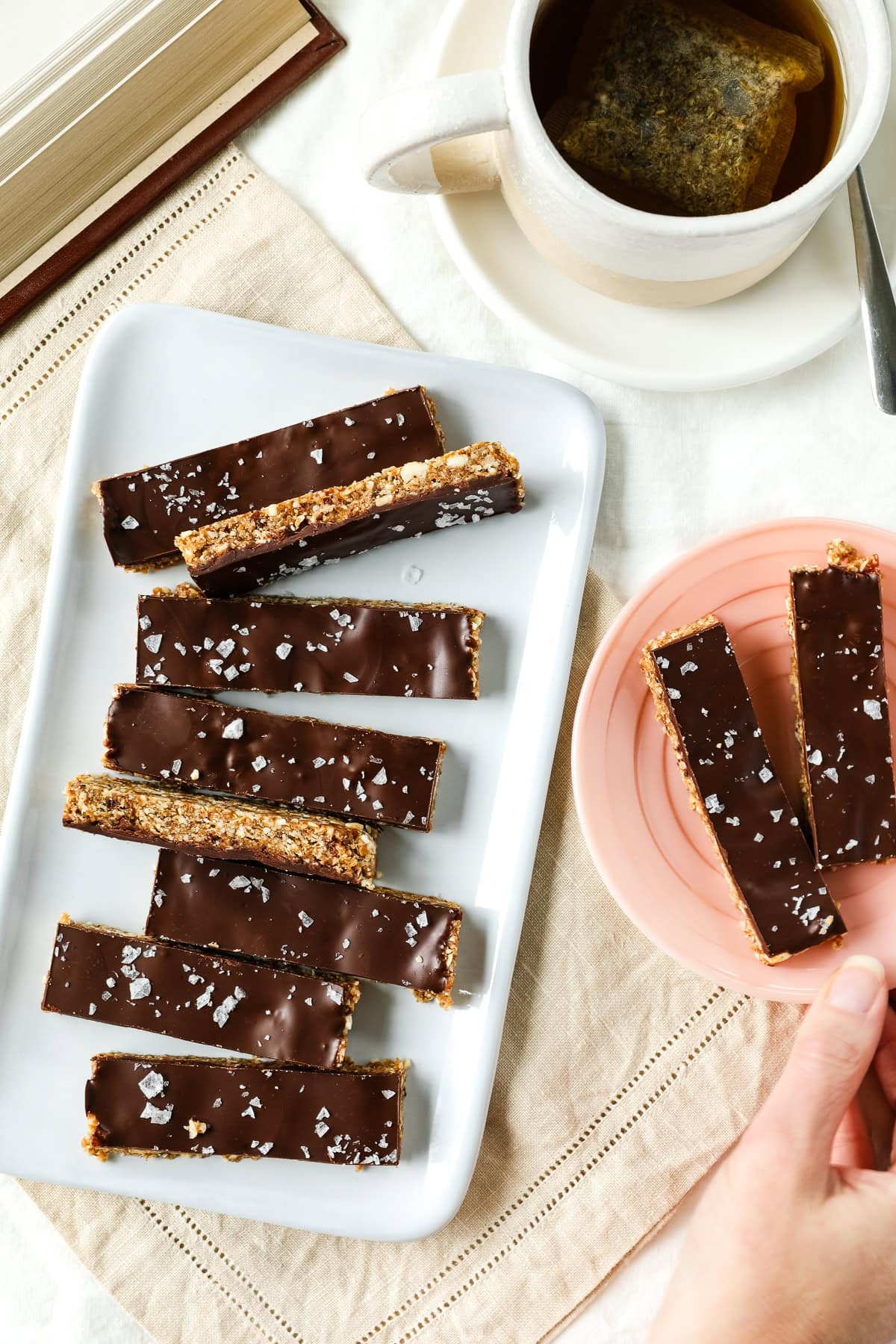 A hand grabs a small plate of date bars next to a larger plate of bars. Tea and a book are nearby.